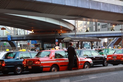 A line of taxis, each with unique color schemes, is parked under a large, elevated roadway in a busy urban setting. A man stands beside one of the taxis, which has a distinct orange stripe pattern. Surrounding infrastructure includes modern buildings with large windows and various signage.