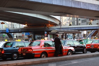 A line of taxis, each with unique color schemes, is parked under a large, elevated roadway in a busy urban setting. A man stands beside one of the taxis, which has a distinct orange stripe pattern. Surrounding infrastructure includes modern buildings with large windows and various signage.