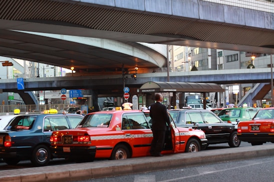 A line of taxis, each with unique color schemes, is parked under a large, elevated roadway in a busy urban setting. A man stands beside one of the taxis, which has a distinct orange stripe pattern. Surrounding infrastructure includes modern buildings with large windows and various signage.