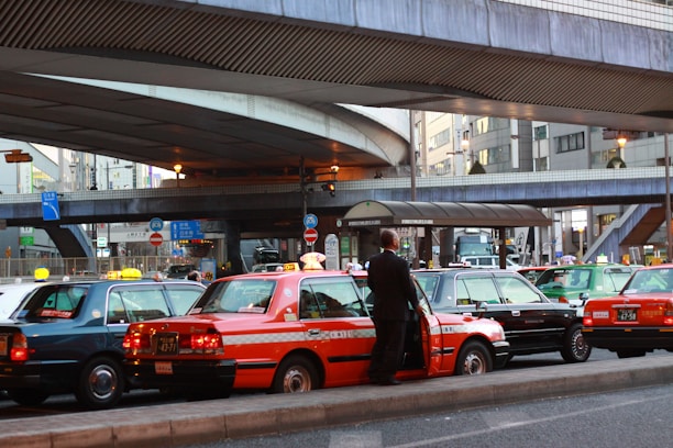 A line of taxis, each with unique color schemes, is parked under a large, elevated roadway in a busy urban setting. A man stands beside one of the taxis, which has a distinct orange stripe pattern. Surrounding infrastructure includes modern buildings with large windows and various signage.
