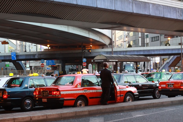 A line of taxis, each with unique color schemes, is parked under a large, elevated roadway in a busy urban setting. A man stands beside one of the taxis, which has a distinct orange stripe pattern. Surrounding infrastructure includes modern buildings with large windows and various signage.