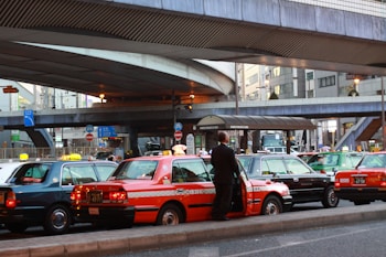 A line of taxis, each with unique color schemes, is parked under a large, elevated roadway in a busy urban setting. A man stands beside one of the taxis, which has a distinct orange stripe pattern. Surrounding infrastructure includes modern buildings with large windows and various signage.
