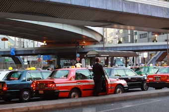 A line of taxis, each with unique color schemes, is parked under a large, elevated roadway in a busy urban setting. A man stands beside one of the taxis, which has a distinct orange stripe pattern. Surrounding infrastructure includes modern buildings with large windows and various signage.