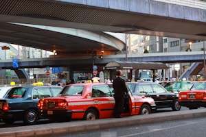 A line of taxis, each with unique color schemes, is parked under a large, elevated roadway in a busy urban setting. A man stands beside one of the taxis, which has a distinct orange stripe pattern. Surrounding infrastructure includes modern buildings with large windows and various signage.