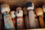 A collection of traditional Japanese Kokeshi dolls made of wood, with simple facial expressions and floral patterns painted on their bodies. The dolls are arranged in a woven basket, showcasing different sizes and designs.
