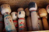 A collection of traditional Japanese Kokeshi dolls made of wood, with simple facial expressions and floral patterns painted on their bodies. The dolls are arranged in a woven basket, showcasing different sizes and designs.