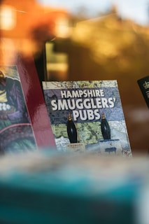 A book with the title 'Hampshire Smugglers' Pubs' is displayed, featuring a map as its background. Bottles of Ringwood Brewery ale are prominently shown on the cover. The setting appears to be a shop with a reflective surface showing vague outlines of the surroundings.