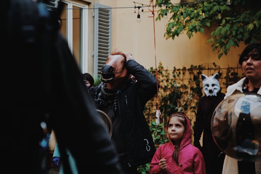 A gathering of people in a yard, some wearing costumes and masks, including a horse and a wolf mask. A child in a pink jacket looks on with curiosity. The setting includes a backdrop of a yellow wall with greenery and string lights overhead.