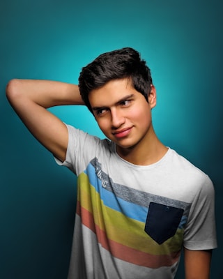 A young person with dark hair is wearing a colorful striped T-shirt. They have one hand behind their head, posing against a smooth teal background. The lighting is focused on their face, giving a vibrant and fresh appearance.