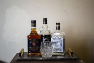 A selection of three liquor bottles arranged on a wooden tray with a crystal whiskey glass placed in front. The bottles include Jim Beam Double Oak, Kraken, and Winchester Bourbon Whiskey. The scene is set against a plain textured wall, giving a classic and refined appearance.
