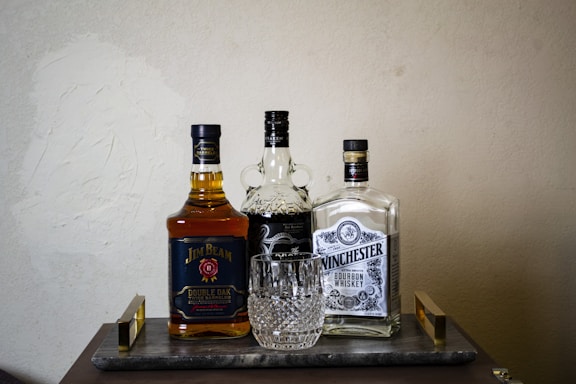 A selection of three liquor bottles arranged on a wooden tray with a crystal whiskey glass placed in front. The bottles include Jim Beam Double Oak, Kraken, and Winchester Bourbon Whiskey. The scene is set against a plain textured wall, giving a classic and refined appearance.