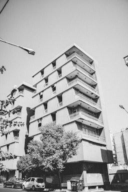 A multi-story apartment building with angular balconies and a striking geometric design. The image captures a few cars parked along the street and a tree providing some greenery in the foreground. A streetlamp arches over the scene, contributing to the urban landscape.