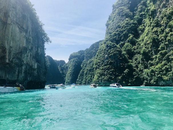 Turquoise waters surrounded by towering limestone cliffs covered with lush green foliage. Several motorboats are traveling through the water, reflecting a serene and vibrant seascape.