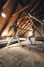 Two dancers perform in an attic with rustic wooden beams and a slanted roof. Light filters through small windows, illuminating their intricate choreography. The dancers display grace and balance amidst the aged, textured setting.