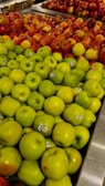 A display of neatly arranged apples in a grocery store, with green apples in the foreground and red apples in the background. Each apple has a small sticker indicating its variety or origin.