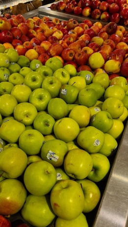 A display of neatly arranged apples in a grocery store, with green apples in the foreground and red apples in the background. Each apple has a small sticker indicating its variety or origin.