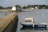 A small boat labeled 'Marguerite' is docked near a stone walkway that extends into a calm body of water. In the background, a quaint house with blue shutters sits on a small island, surrounded by greenery and additional houses further away. The sky is overcast, casting a soft light over the scene.