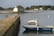 A small boat labeled 'Marguerite' is docked near a stone walkway that extends into a calm body of water. In the background, a quaint house with blue shutters sits on a small island, surrounded by greenery and additional houses further away. The sky is overcast, casting a soft light over the scene.