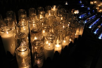 A collection of lit candles in tall glass holders emits a warm glow, creating a serene and contemplative atmosphere. The arrangement appears orderly, with flickering flames casting soft shadows and reflections on the surrounding glass surfaces.