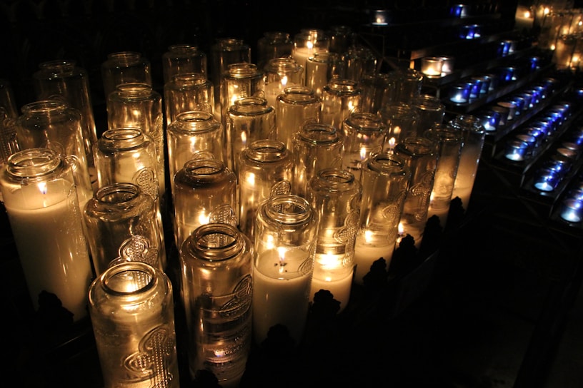 A collection of lit candles in tall glass holders emits a warm glow, creating a serene and contemplative atmosphere. The arrangement appears orderly, with flickering flames casting soft shadows and reflections on the surrounding glass surfaces.