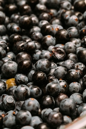 A close-up view of a pile of dark purple berries, densely packed together with one or two green berries interspersed among them. The berries have a shiny, smooth surface and some show water droplets, indicating freshness.