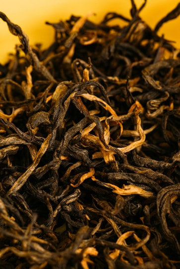 A close-up view of loose, dried black tea leaves with a combination of dark and light brown hues, set against a soft yellow background. The leaves have a twisted, curly texture.