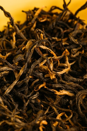 A close-up view of loose, dried black tea leaves with a combination of dark and light brown hues, set against a soft yellow background. The leaves have a twisted, curly texture.