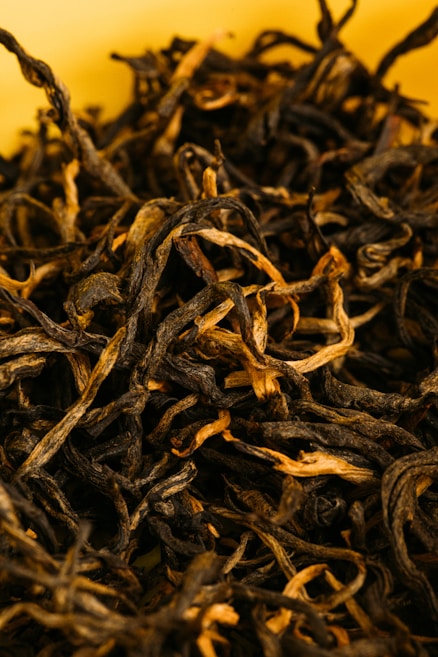 A close-up view of loose, dried black tea leaves with a combination of dark and light brown hues, set against a soft yellow background. The leaves have a twisted, curly texture.