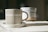 A ceramic mug with a vertical striped pattern sits on a matching saucer. The mug has an earthy beige tone, and in the background, there is a blurred out similar ceramic item. Both items are placed on a fabric surface that seems to be softly lit by natural light.