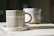 A ceramic mug with a vertical striped pattern sits on a matching saucer. The mug has an earthy beige tone, and in the background, there is a blurred out similar ceramic item. Both items are placed on a fabric surface that seems to be softly lit by natural light.