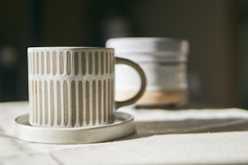 A ceramic mug with a vertical striped pattern sits on a matching saucer. The mug has an earthy beige tone, and in the background, there is a blurred out similar ceramic item. Both items are placed on a fabric surface that seems to be softly lit by natural light.
