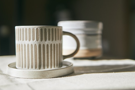 A ceramic mug with a vertical striped pattern sits on a matching saucer. The mug has an earthy beige tone, and in the background, there is a blurred out similar ceramic item. Both items are placed on a fabric surface that seems to be softly lit by natural light.