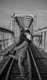 A couple embraces romantically on a railway track, with an old metal bridge in the background. The image is black and white, capturing an intimate moment as one person lifts the other. The bridge and tracks create a strong sense of depth.