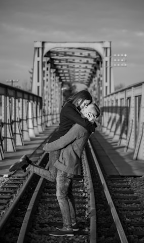 A couple embraces romantically on a railway track, with an old metal bridge in the background. The image is black and white, capturing an intimate moment as one person lifts the other. The bridge and tracks create a strong sense of depth.