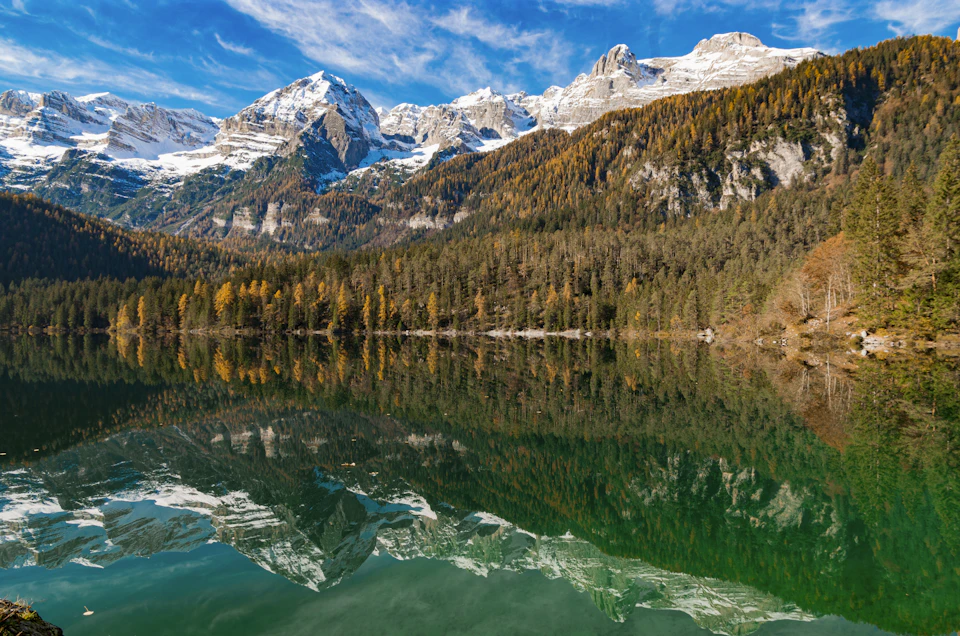 The image depicts a serene mountain lake surrounded by lush green trees and snow-capped mountains. The calm, reflective surface of the lake mirrors the surrounding landscape, creating a beautiful symmetry. The mountains in the background are covered in snow, adding depth to the scene. The sky above is clear blue with scattered clouds, providing a perfect backdrop for this picturesque view.