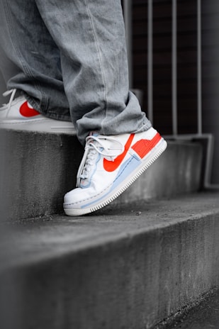 A person wearing light-colored denim jeans stands on a concrete stair, showcasing a pair of stylish white sneakers with vibrant orange accents and a blue design element. The scene is urban with metal railings in the background.