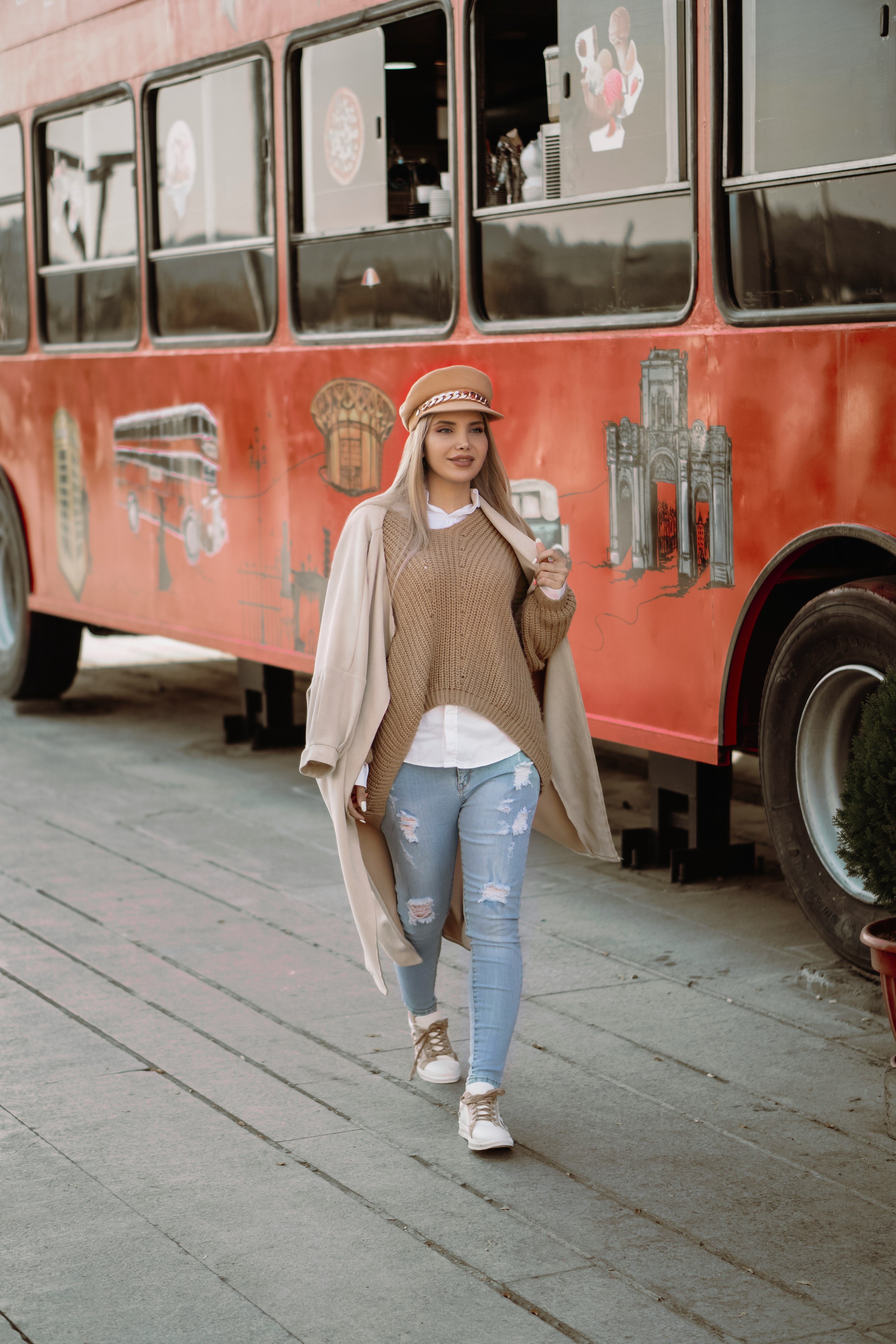 woman in white knit sweater and blue denim jeans standing beside red bus