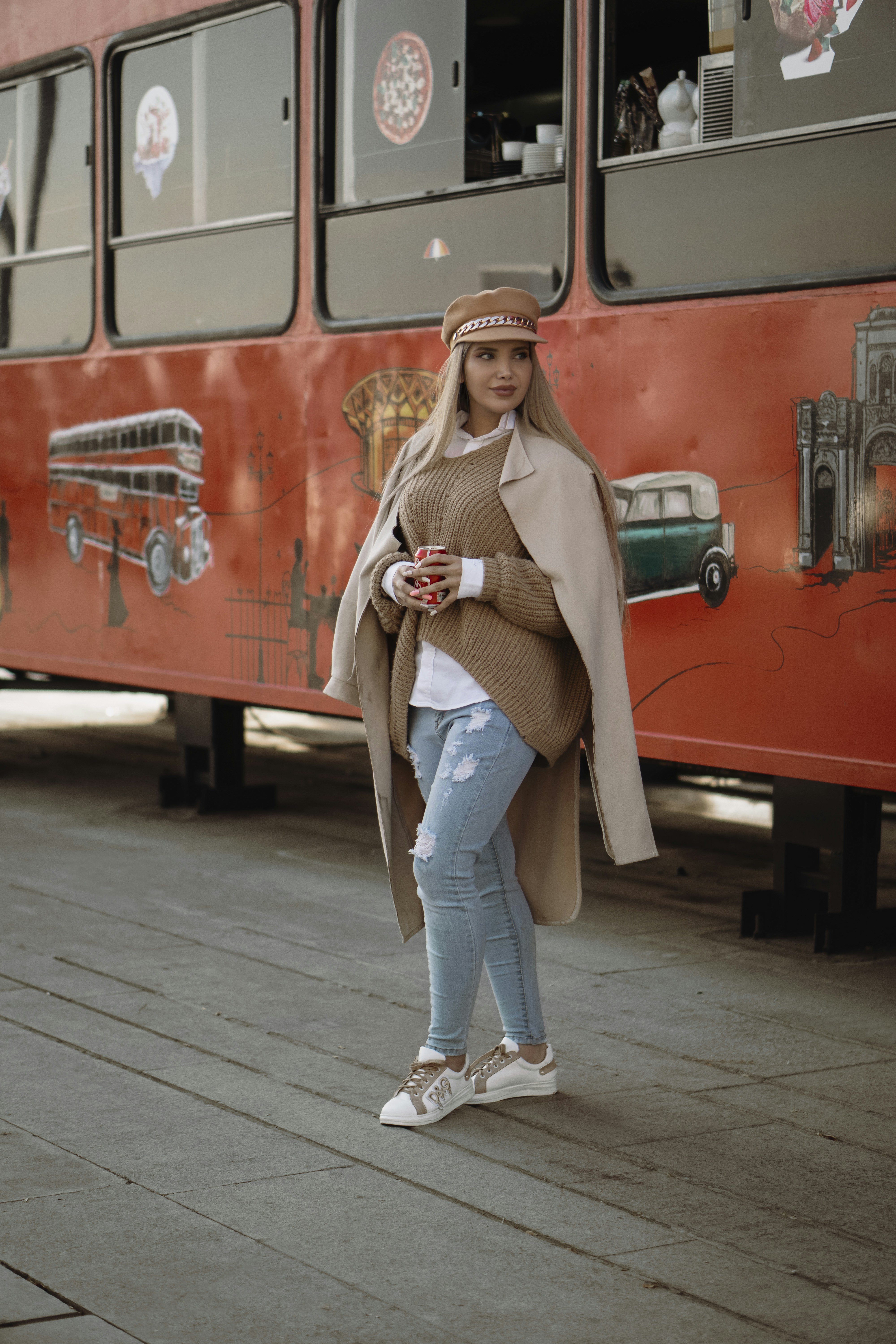 Wanita dengan mantel coklat dan jeans denim biru berdiri di samping bus merah