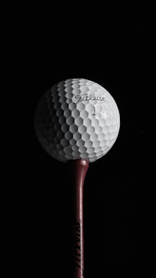 A close-up view of a golf ball positioned on a tee against a dark background. The golf ball displays a dimpled texture and has the brand name 'Titleist' along with the number '1' printed on it.