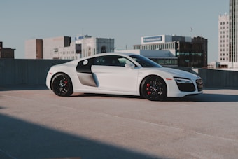 A sleek, white sports car is parked on a rooftop with a cityscape in the background. The car has a smooth, aerodynamic design with noticeable black accents on its sides and wheels. The building in the background suggests an urban environment with clear skies overhead.