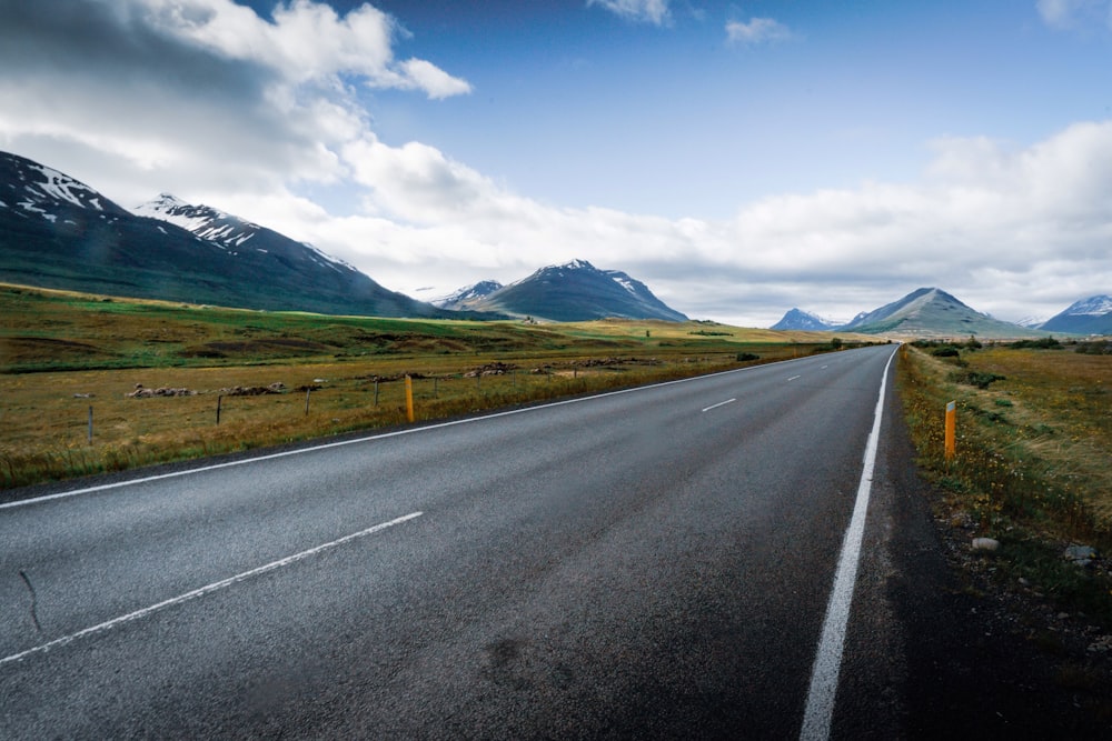 gray concrete road near green grass field and mountains under blue sky and white clouds during