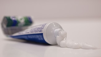 A tube of toothpaste lies on a flat surface with its cap off. A spiraled dollop of white toothpaste is squeezed out onto the surface next to the tube, which has a blue, green, and white design.