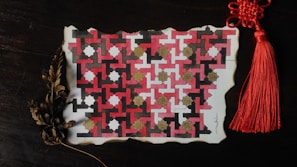 A piece of paper features an intricate geometric pattern with red, black, white, and gold shapes interlocking in a repetitive design. The paper's edges appear to be slightly burnt, giving it an aged, vintage look. A red tassel with a complex knot is attached to its top right corner. Below, dried brown leaves add an organic element to the scene.