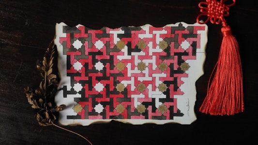 A piece of paper features an intricate geometric pattern with red, black, white, and gold shapes interlocking in a repetitive design. The paper's edges appear to be slightly burnt, giving it an aged, vintage look. A red tassel with a complex knot is attached to its top right corner. Below, dried brown leaves add an organic element to the scene.