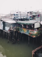 A cluster of stilt houses extends over a body of water. The structures are constructed with a variety of materials, including corrugated metal, wood, and tarpaulin. A small balcony area is adorned with potted plants and a bright yellow chair. Clothes hang from a line, suggesting this area is inhabited. Several makeshift ladders provide access between levels.
