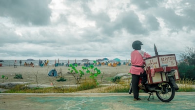 A person wearing a helmet and a pink jacket stands next to an ice cream motorcycle on a beach. The motorcycle has a large insulated box labeled 'Es Potong' on the back. In the background, people are gathered on the sandy beach, with some sitting under colorful umbrellas. The sky is overcast with heavy clouds, creating a moody atmosphere.