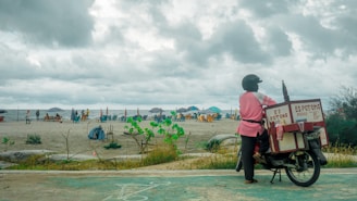 A person wearing a helmet and a pink jacket stands next to an ice cream motorcycle on a beach. The motorcycle has a large insulated box labeled 'Es Potong' on the back. In the background, people are gathered on the sandy beach, with some sitting under colorful umbrellas. The sky is overcast with heavy clouds, creating a moody atmosphere.