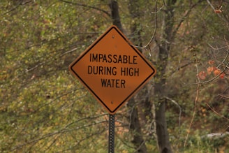 A yellow warning sign in a wooded area contains the text 'IMPASSABLE DURING HIGH WATER'. The background shows trees with a mix of green and autumn-colored leaves.