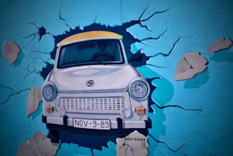 A mural depicting a car breaking through a painted wall, creating the illusion of a crash. The car is in the foreground with its headlights and bumper visibly prominent. The background is a vibrant blue wall with cracks and debris painted around the car, enhancing the effect of it bursting through.