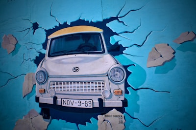A mural depicting a car breaking through a painted wall, creating the illusion of a crash. The car is in the foreground with its headlights and bumper visibly prominent. The background is a vibrant blue wall with cracks and debris painted around the car, enhancing the effect of it bursting through.
